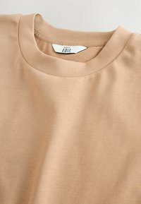 Camiseta de algodón beige de cuello redondo con una textura suave, cuello acanalado y detalles de costura sutiles. La etiqueta dice "NEXT EDIT."