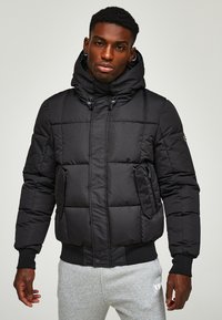 Schwarze Pufferjacke mit Kapuze, ausgestattet mit einem gesteppten Design, Reißverschlusstaschen und gerippten Bündchen. Der Stoff wirkt glatt und leicht.
