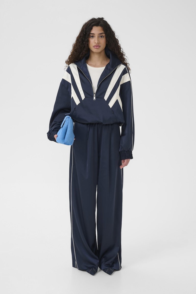 Veste sportive bleu marine avec motif chevron blanc, poignets élastiques, associée à un pantalon ample assorti et une petite pochette serviette bleue.