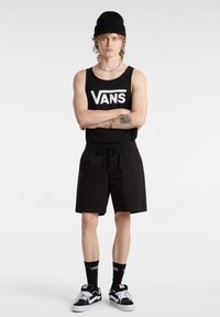 Canotta nera con logo "VANS", pantaloni corti neri con cordoncino, calze nere con logo e scarpe nere e bianche. Il modello è in piedi di fronte.