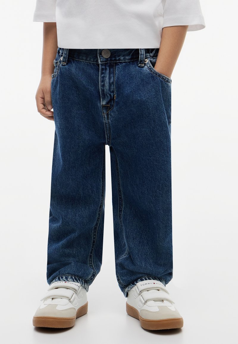 Enfant portant un jean bleu foncé ample et des baskets blanches avec semelles marron, debout les mains dans les poches sur un fond uni.