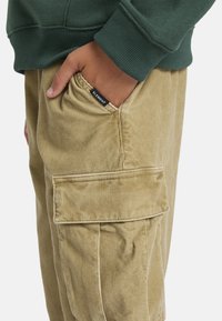 Beige Corduroy-Cargohosen mit Taschen und einem kleinen schwarzen Etikett. Getragen mit einem grünen Sweatshirt, das die strukturierten Stoffdetails betont.