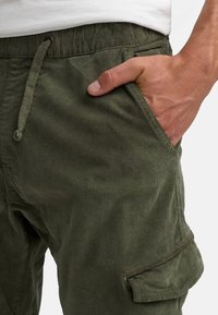 Pantaloni cargo verde oliva con vita regolabile, tasca laterale e tessuto texture. Presentano una vestibilità casual e rilassata e dettagli di cucitura.