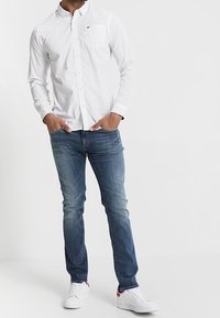 Camisa blanca de manga larga con botones y un bolsillo, combinada con jeans de mezclilla azul. Los zapatos son blancos con acentos rojos, de pie frente a un fondo liso.