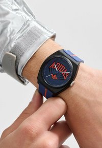Montre noire avec un cadran bleu arborant un logo "Adidas", un bracelet texturé bleu et rouge, et un accent argenté sur le côté.