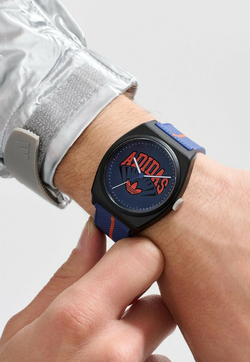 Montre noire avec un cadran bleu arborant un logo "Adidas", un bracelet texturé bleu et rouge, et un accent argenté sur le côté.