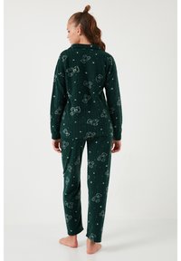 Femeie stând desculță, purtând un set de pijama verde închis cu mâneci lungi cu modele de ursuleț și stele, văzută din spate.