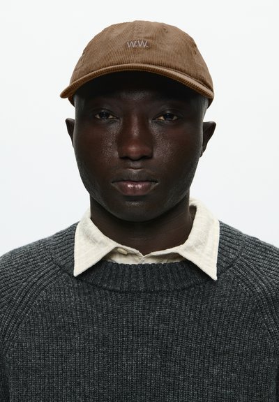 Barna kordbársony sapka "W.W." hímzéssel, amelyet egy texturált sötétszürke pulóverrel és alatta egy világosszürke kockás blúzzal párosítottak.