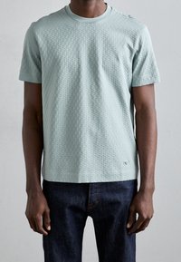 Mátově zelené tričko s krátkým rukávem, které má texturovaný vaflový vzor a kulatý výstřih. Tričko má viditelné švy a malé logo na lemu.