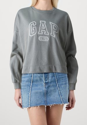 Vrouw draagt een grijze GAP-trui met de tekst "1969" en een blauwe denim minirok met rafelige zoom, staand tegen een effen achtergrond.