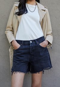 Chaqueta de moto de cuero beige sobre una camiseta blanca, combinada con shorts de mezclilla oscura con dobladillos deshilachados y bolsillos frontales.