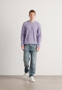 Vyrų modelis vilkintis šviesiai violetinės spalvos ilgomis rankovėmis marškiniais, suplyšusiais mėlynais džinsais ir baltais sportbačiais, stovi prieš neutralų foną.