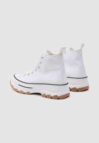 Zapatillas altas blancas de lona con suela de goma texturizada, que presentan una franja negra y ojales plateados. Suela exterior marrón claro.