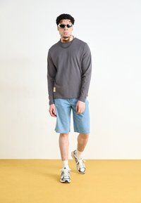 Gray long-sleeve shirt, denim shorts, and athletic shoes. Sunglasses and a beaded necklace complete the look, against a light background.