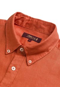 Camicia di lino color ruggine con colletto a camicia, bottoni bianchi e un'etichetta viola con testo dorato. Texture liscia e design casual.