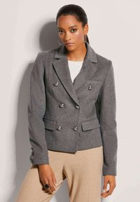 Grau Wollblazer mit doppelter Knopfleiste, silbernen Knöpfen, Reverskragen und Vordertaschen. Kombiniert mit beigen Hosen.