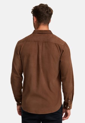 Man met kort bruin haar die een lange mouwen bruine blouse met knopen draagt, gefotografeerd van achteren tegen een effen achtergrond.