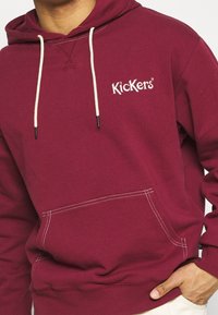 Mann trägt ein weinrotes Kapuzenshirt mit weißen Kordeln und "Kickers"-Logo auf der Brust, die Hand in der vorderen Tasche.