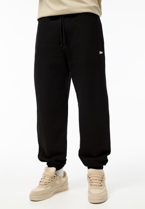 Zwarte katoenen sweatpants met een elastische tailleband en boorden, voorzien van een klein wit logo aan de linkerkant; gecombineerd met lichtbeige sneakers.