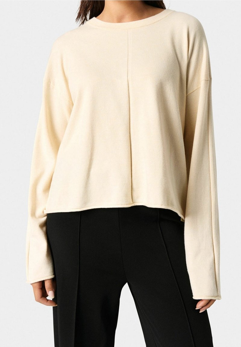 Femme portant un pull crème ample à manches longues avec une couture centrale, associé à un pantalon noir taille haute sur un fond uni.