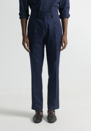 Pantalones azul marino de corte ajustado con cintura alta, pliegues y textura suave. Combinados con zapatos oscuros trenzados, sobre un fondo liso.