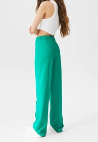 Femme aux cheveux longs portant un crop top blanc et un pantalon large taille haute vert, debout les bras croisés sur fond blanc.