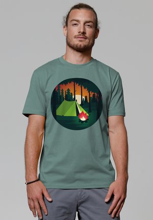Groene katoen T-shirt met een geïllustreerd cirkelontwerp met een tent, kampvuur en bos, tegen een oranje zonovergoten achtergrond.