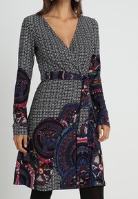 Langärmeliges Wickelkleid aus dunklem Stoff mit geometrischen Mustern in Schwarz und Weiß, versehen mit bunten kreisförmigen Akzenten am Rock.