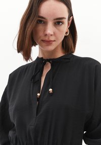 Schwarzes Stoffkleid mit Stehkragen und Bindebandverschluss, verziert mit Holzkugeln. Das Design ist entspannt und leicht texturiert.
