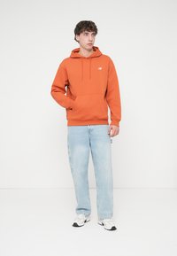 Πορτοκαλί φούτερ με κουκούλα και τσέπη μπροστά, συνδυασμένο με ανοιχτό μπλε τζιν και λευκά αθλητικά παπούτσια. Διακριτική λεπτομέρεια με λογότυπο New Balance.