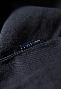 Dunkelblaue Denim-Stoff mit einer dezenten Textur, versehen mit einem sichtbaren blauen Etikett, auf dem "LINDBERGH" in weißer Schrift steht.