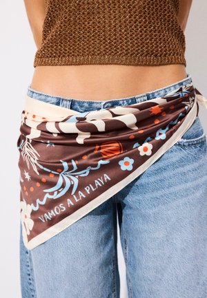 Braun gemustertes Tuch mit Blumen und dem Text „VAMOS A LA PLAYA“, um die Taille gebunden über blauen Jeans und einem braunen Strick-Cropped-Top.