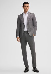Un costume gris en tissu texturé avec une coupe ajustée, comprenant une veste à un bouton et un pantalon slim, associé à une chemise blanche.