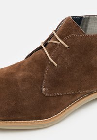 Ruskeat nubuk-nilkkurit, joissa on sileä pinta, beige kengännauhat ja tikattu pohja. Muotoilussa on pyöristetty kärki ja matala korko.