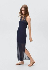 Abito maxi senza maniche blu navy con uno spacco alto, realizzato in materiale leggero. Abbinato a sandali marroni con cinturini. Texture liscia e design semplice.