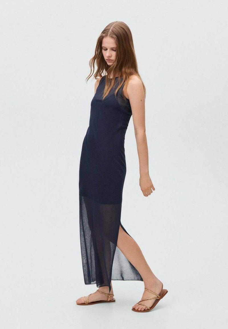 Abito maxi senza maniche blu navy con uno spacco alto, realizzato in materiale leggero. Abbinato a sandali marroni con cinturini. Texture liscia e design semplice.
