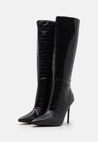 Svarta högklackade stövlar med glansig krokodiltextur, spetsig tå och en åtsittande design som går upp till knäna. Stiliga klackar bidrar med extra höjd.