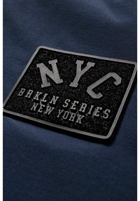 Patch rectangulaire noir avec l’inscription « NYC BRKLN SERIES NEW YORK » en lettres grasses sur fond de tissu bleu foncé.