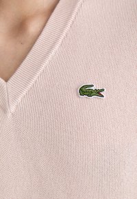 Pull en tricot rose clair avec un col en V, arborant un logo de crocodile vert brodé sur la poitrine gauche.