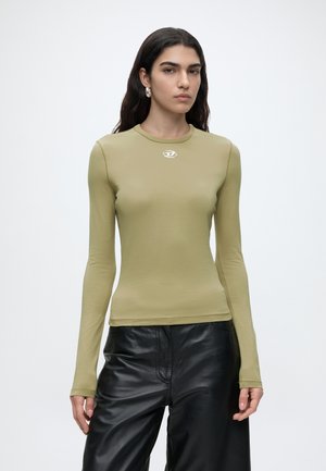 Femme aux longs cheveux foncés portant des boucles d’oreilles en créoles, un haut moulant à manches longues vert olive et un pantalon en cuir noir ample sur un fond uni.