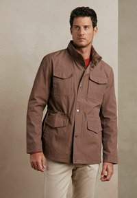 Brązowa, wodoodporna kurtka z podniesionym kołnierzem, czterema przednimi kieszeniami, ściągaczem w pasie i zatrzaskami. Gładka tekstura, casualowy styl.