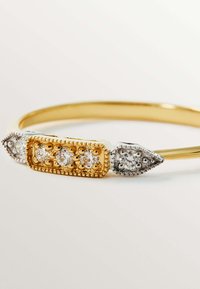 Brazalete de oro y plata con acentos de gemas; cuenta con un centro rectangular de oro y dos extremos puntiagudos, cada uno adornado con piedras claras.