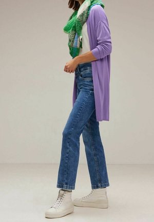 Femme portant un jean bleu, des baskets blanches montantes, un cardigan lavande, une chemise blanche et une écharpe à carreaux verts, debout devant un fond uni.