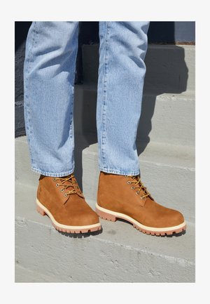 Bruine suède enkellaarsjes met gele veters en witte zolen, met een robuust loopvlak, gedragen met lichtblauwe denim jeans op trappen.