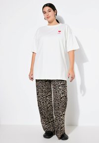 T-shirt bianco oversize con un cuore rosso e il testo "TU," abbinato a pantaloni larghi con stampa leopardata beige e nera e scarpe nere.