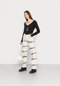 Body preto de manga longa com detalhe em forma de chave, calças de cintura alta tie-dye em marfim e cinza, e botas de couro pretas com atacadores. Mala branca.