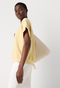Světle žlutý oversized top s krátkými rukávy, doplněný bílými kalhotami. Velká neutrální kabelka leží na boku modelky.