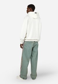 Weißer Kapuzenpullover mit Kordelzug, kombiniert mit übergroßen hellblauen Jeans mit Seitenstreifen. Modell steht vor einem schlichten Hintergrund.