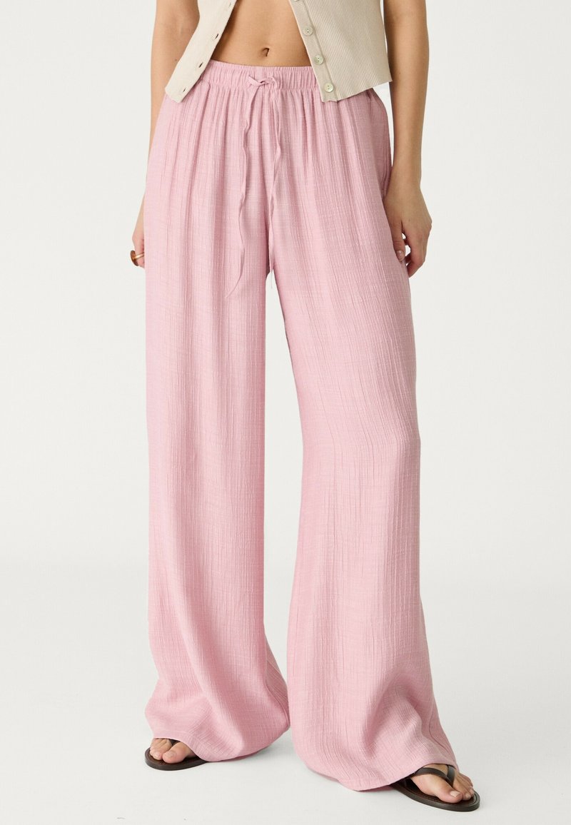 Femme portant un pantalon ample rose clair texturé à jambes larges avec une ceinture élastique et un cordon, associé à des tongs noires.