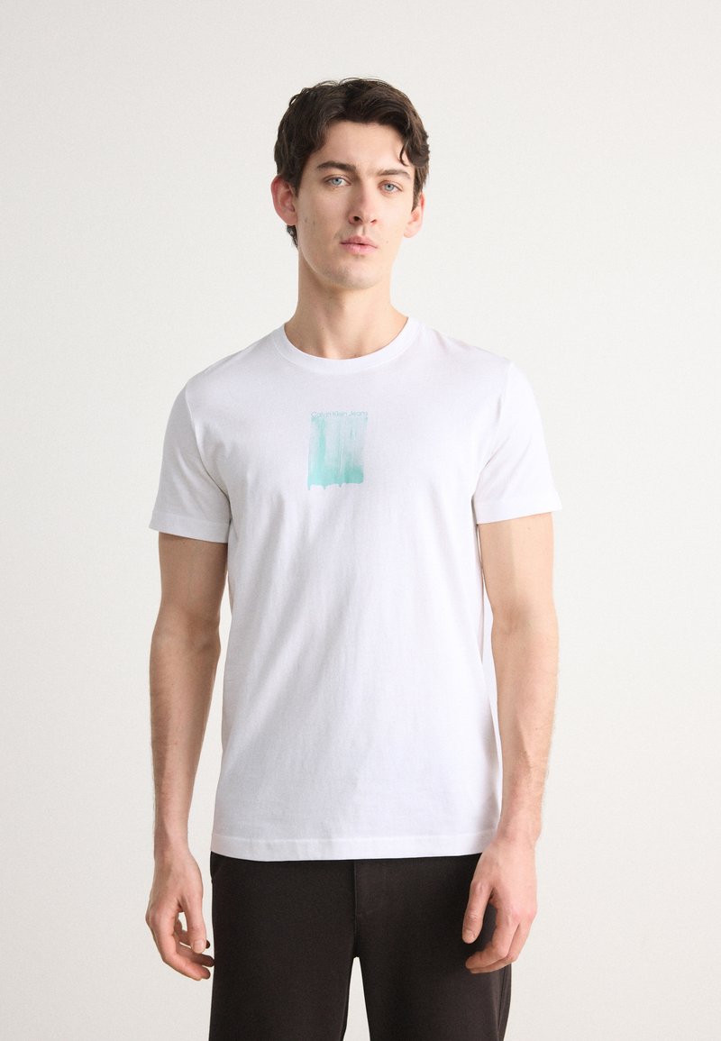 Camiseta blanca de algodón de manga corta con un diseño de pincelada azul claro en el pecho, modelo vistiendo pantalones oscuros, fondo neutral.
